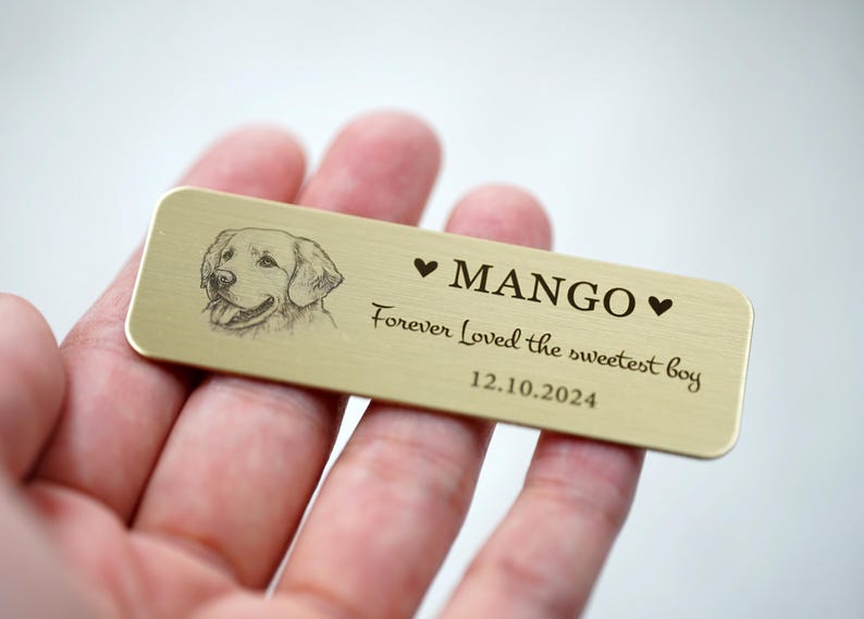 May include: A rectangular, gold-colored metal tag with a sketch of a dog's face. The tag reads "MANGO" with a heart symbol on each side, and the text "Forever Loved the sweetest boy 12.10.2024".