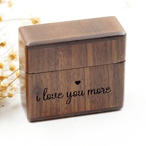 Peut inclure: Une petite bo&icirc;te en bois avec un couvercle &agrave; charni&egrave;re. La bo&icirc;te est de couleur brun fonc&eacute; et porte les mots "i love you more" grav&eacute;s sur le devant, ainsi qu'un petit c&oelig;ur.
