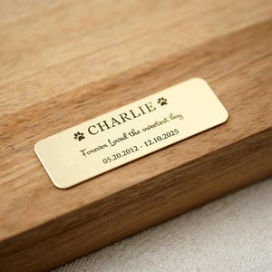 May include: A rectangular gold-colored plaque with rounded corners, engraved with the name "CHARLIE", paw prints, and the phrase "Forever Loved the sweetest boy". The dates 05.20.2012 - 12.10.2025 are also inscribed. The plaque is set on a wooden surface.