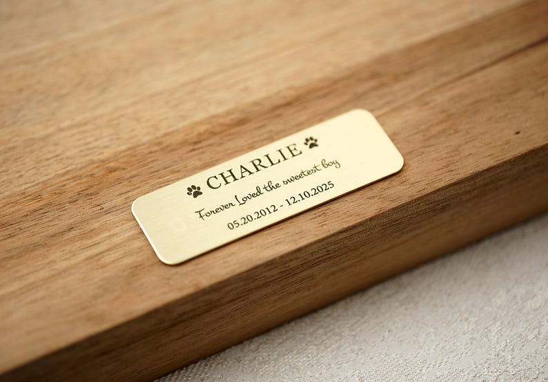 May include: A rectangular gold-colored plaque with rounded corners, engraved with the name "CHARLIE", paw prints, and the phrase "Forever Loved the sweetest boy". The dates 05.20.2012 - 12.10.2025 are also inscribed. The plaque is set on a wooden surface.