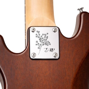 May include: Close-up of the back of a brown electric guitar. A silver plate with a floral design and the initials "SW" is attached to the guitar's body. The guitar neck is visible, with a light wood finish.