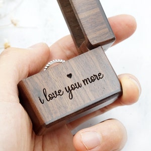 Peut inclure: Une bo&icirc;te &agrave; bague en bois avec l'inscription "i love you more" et un petit c&oelig;ur. La bo&icirc;te est ouverte et une bague en argent se trouve &agrave; l'int&eacute;rieur.
