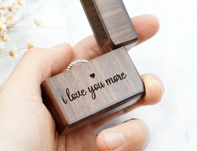 Peut inclure: Une bo&icirc;te &agrave; bague en bois avec l'inscription "i love you more" et un petit c&oelig;ur. La bo&icirc;te est ouverte et une bague en argent se trouve &agrave; l'int&eacute;rieur.