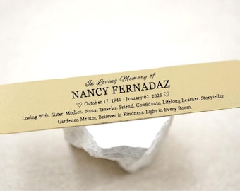 May include: A rectangular, light tan memorial plaque with rounded corners, bearing the inscription "In Loving Memory of NANCY FERNADAZ." The text includes dates, and a list of loving attributes. The plaque rests on a white, textured surface.