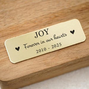May include: A rectangular gold-colored plaque with rounded corners, engraved with the word "JOY", the phrase "Forever in our hearts", and the years "2010 - 2025". Two black heart symbols flank the text. The plaque is set on a wooden surface.