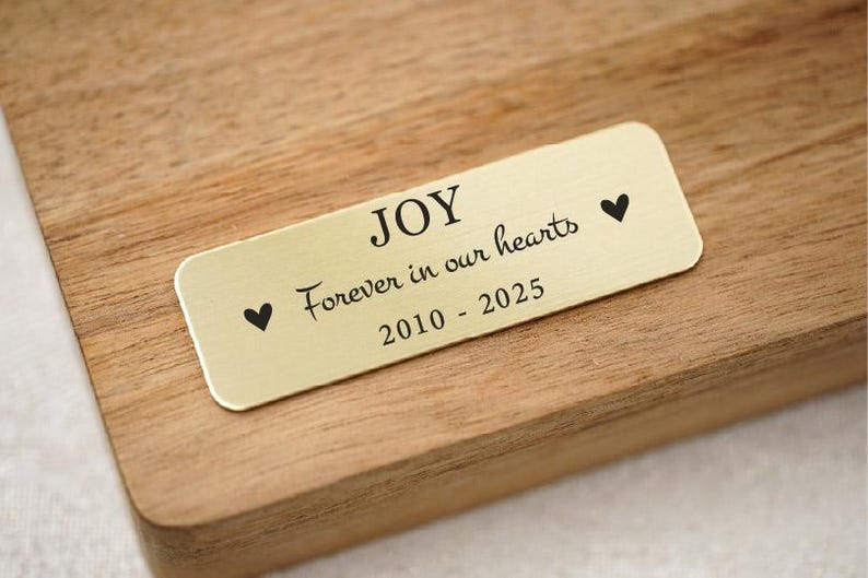 May include: A rectangular gold-colored plaque with rounded corners, engraved with the word "JOY", the phrase "Forever in our hearts", and the years "2010 - 2025". Two black heart symbols flank the text. The plaque is set on a wooden surface.