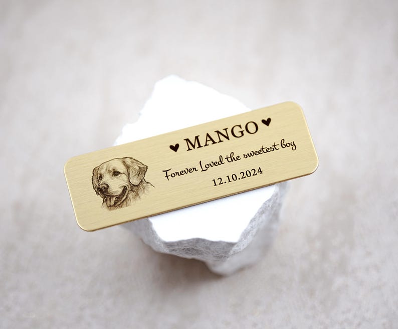 May include: A rectangular, gold-colored metal tag with a detailed engraving of a dog's face on the left side. The tag has the name "MANGO" with a heart symbol above it, and the text "Forever Loved the sweetest boy 12.10.2024".