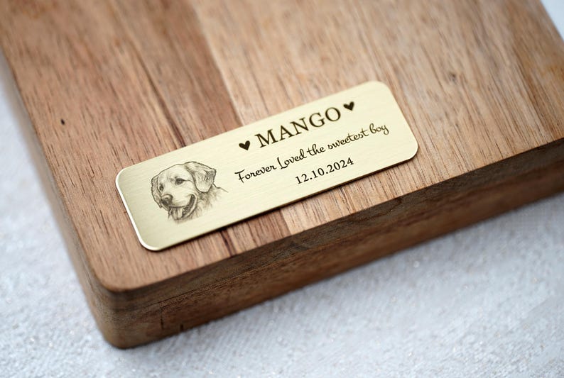 May include: A rectangular, gold-colored metal plaque with rounded corners, engraved with the name "MANGO" and a dog portrait. Text reads "Forever Loved the sweetest boy 12.10.2024". The plaque is set on a wooden surface.