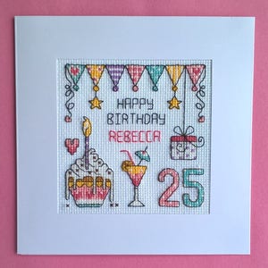 May include: Cross-stitch birthday card with a white mount. The design includes a cupcake with a lit candle, a cocktail, a gift, and the number 25. The words "HAPPY BIRTHDAY REBECCA" are stitched across the top.