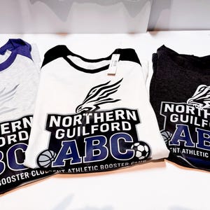 May include: Three athletic booster club t-shirts. The shirts are white, gray, and dark gray, each with a different color scheme. The shirts feature the text "Northern Guilford ABC" with a stylized eagle graphic and sports ball icons.