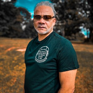 May include: A man wearing a dark green t-shirt with the text "THE WORLD NEEDS WHO YOU WERE MADE TO BE" and sunglasses. The background is a blurred outdoor scene with trees and a blue sky.