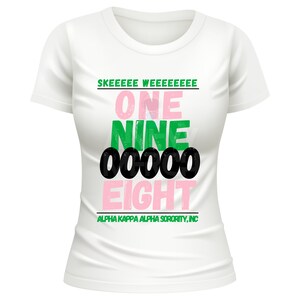 May include: White t-shirt with a crew neck featuring text in pink, green, and black. The text includes "SKEEEEEE WEEEEEEEEE", "ONE NINE 00000 EIGHT", and "ALPHA KAPPA ALPHA SORORITY, INC."