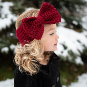 May include: A close-up of a child wearing a large, handmade, burgundy crochet bow headband. The bow is centered on the head and is a prominent feature. The background is a snowy, outdoor scene.