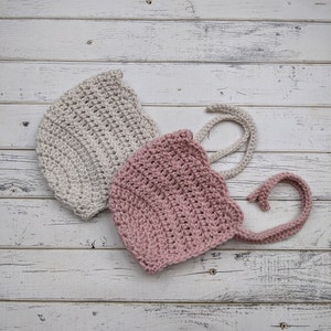 May include: Two crocheted baby bonnets, one in beige and one in pink, with ties. The bonnets are on a white wooden background.