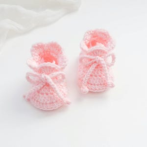 May include: A pair of light pink baby booties crafted with a crochet technique. Each bootie features a ruffled top edge and a matching pink yarn tie threaded through the ankle area, tied into a bow at the front of the footwear.