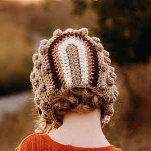 May include: A handmade crochet hat in shades of brown, beige, and white. The hat features a textured, bobble stitch design around the brim and a striped pattern across the crown. The hat is shown from the back, with a person wearing an orange shirt.