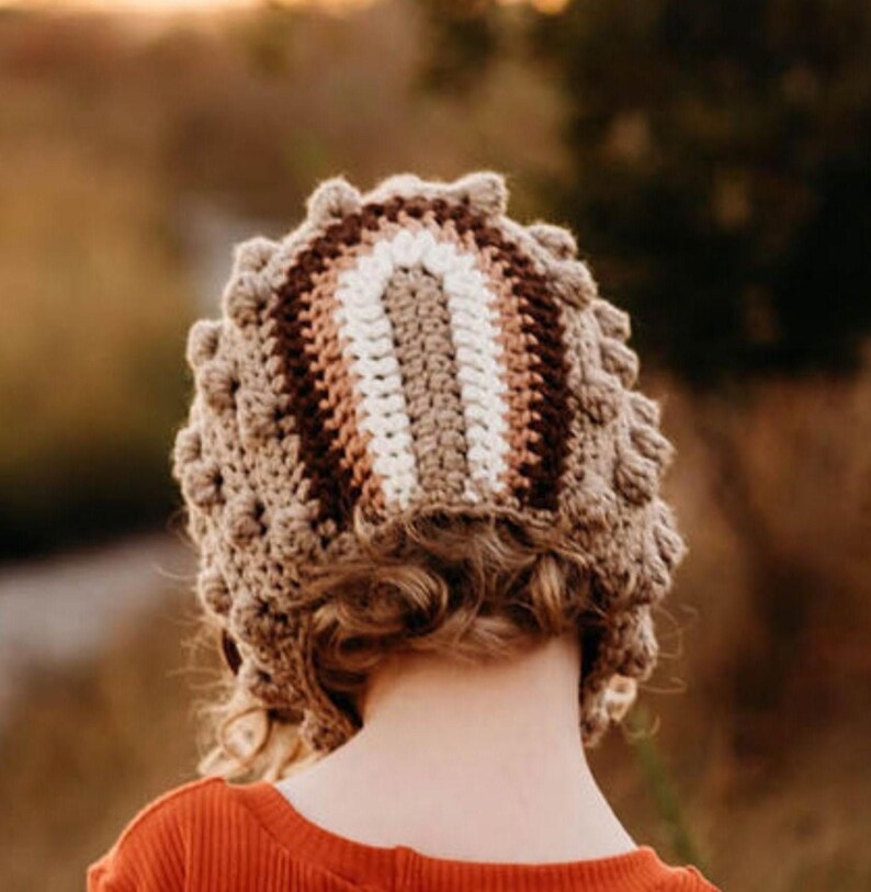 May include: A handmade crochet hat in shades of brown, beige, and white. The hat features a textured, bobble stitch design around the brim and a striped pattern across the crown. The hat is shown from the back, with a person wearing an orange shirt.