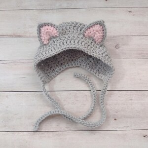 May include: A light gray crochet hat featuring two pointed cat ears with soft pink inner detailing. The hat is constructed with a textured yarn stitch and includes two long, matching gray braided ties extending from the bottom edges.