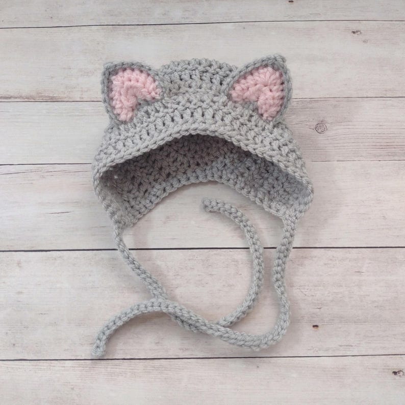 May include: A light gray crochet hat featuring two pointed cat ears with soft pink inner detailing. The hat is constructed with a textured yarn stitch and includes two long, matching gray braided ties extending from the bottom edges.