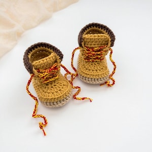 May include: A pair of tan crochet baby booties designed to resemble work boots. Each boot features a dark brown cuff, a light tan sole, and red and yellow braided laces that hang down the sides of the footwear.