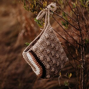 May include: A handmade crochet bag in shades of brown, beige, and white, hanging from a tree branch. The bag features a textured, bobble stitch design and a striped pattern near the opening. The bag is suspended by a knotted cord.