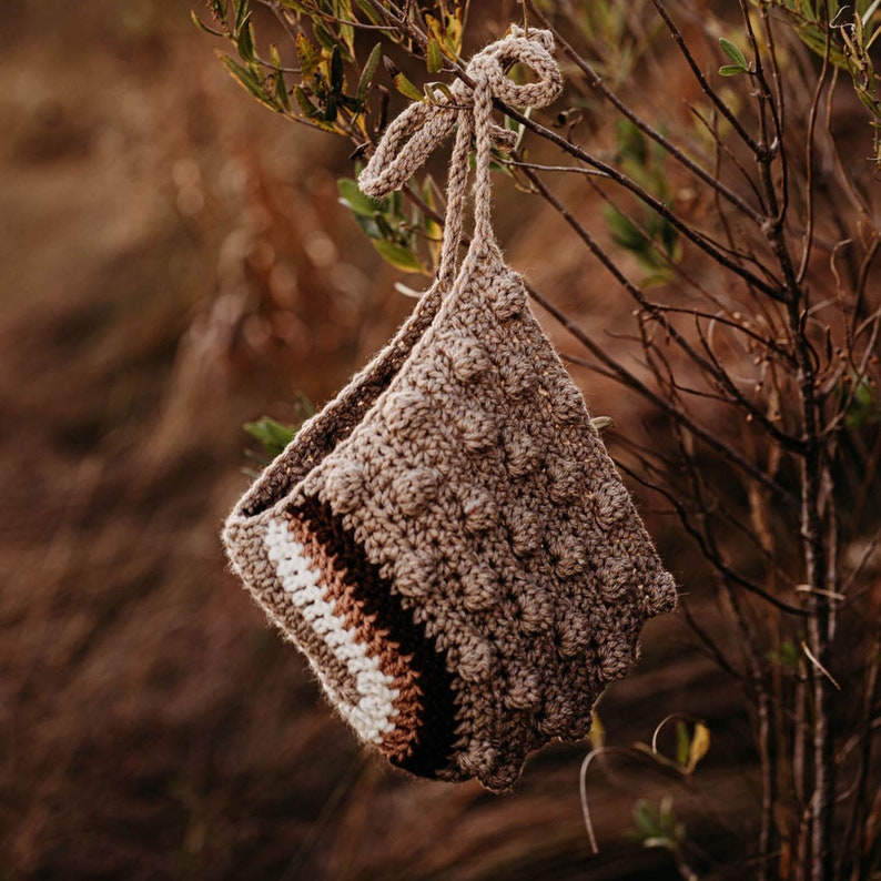 May include: A handmade crochet bag in shades of brown, beige, and white, hanging from a tree branch. The bag features a textured, bobble stitch design and a striped pattern near the opening. The bag is suspended by a knotted cord.