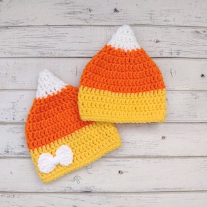 May include: Two handmade crochet hats in the shape of candy corn. The hats feature a white tip, an orange middle, and a yellow base. One hat has a small white bow attached to the yellow base. The hats are displayed on a white wooden surface.