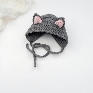 May include: A dark gray crocheted hat featuring two pointed cat ears with light pink centers. The hat is constructed with a textured yarn stitch and includes two long crocheted ties extending from the sides to secure the item under the chin.