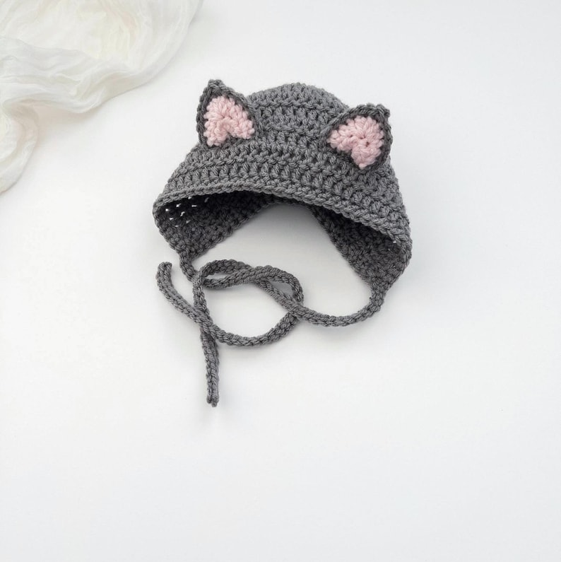 May include: A dark gray crocheted hat featuring two pointed cat ears with light pink centers. The hat is constructed with a textured yarn stitch and includes two long crocheted ties extending from the sides to secure the item under the chin.