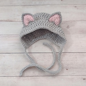 May include: A light gray crochet hat featuring two pointed cat ears with soft pink inner detailing. The hat is constructed with a textured yarn stitch and includes two long, matching gray braided ties extending from the bottom edges.