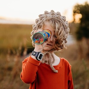 May include: A child wearing a rust-colored long-sleeved shirt and a crocheted bonnet looks through a colorful kaleidoscope. The kaleidoscope is cylindrical with a blue and yellow design and filled with colorful beads. A black and white watch is on the child's wrist.