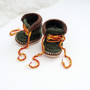 May include: A pair of handmade crochet baby boots in dark olive green with brown cuffs. The boots feature bright yellow and red braided laces that extend outward. The soles are a light beige color, and the overall construction shows a textured yarn stitch pattern throughout the footwear.