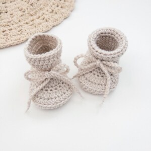 May include: A pair of light beige crochet baby booties with delicate bow ties. The booties are handmade with a textured stitch, and a matching crocheted hat is partially visible in the background. The soft, neutral colors suggest a baby item.