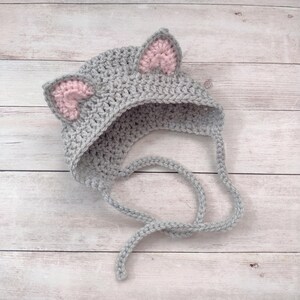 May include: A gray crocheted bonnet featuring two pointed ears with light pink inner details. The hat is constructed from a textured yarn stitch and includes two long, matching gray crocheted ties extending from the base to secure the item.
