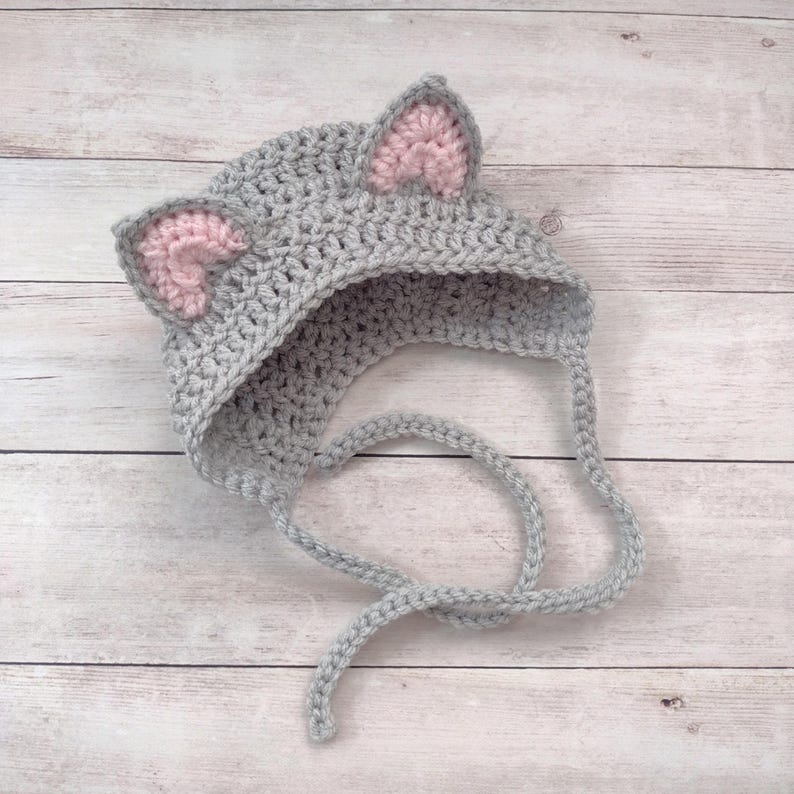 May include: A gray crocheted bonnet featuring two pointed ears with light pink inner details. The hat is constructed from a textured yarn stitch and includes two long, matching gray crocheted ties extending from the base to secure the item.