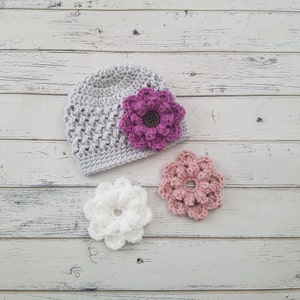 May include: A gray crocheted hat with a pink flower and two smaller white flowers. The hat and flowers are on a white wooden background.