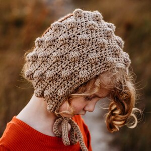 May include: A tan crocheted bonnet with a textured, bobble stitch design. The bonnet has a rounded shape and ties under the chin with a matching tan cord. The bonnet is worn by a person wearing an orange shirt.