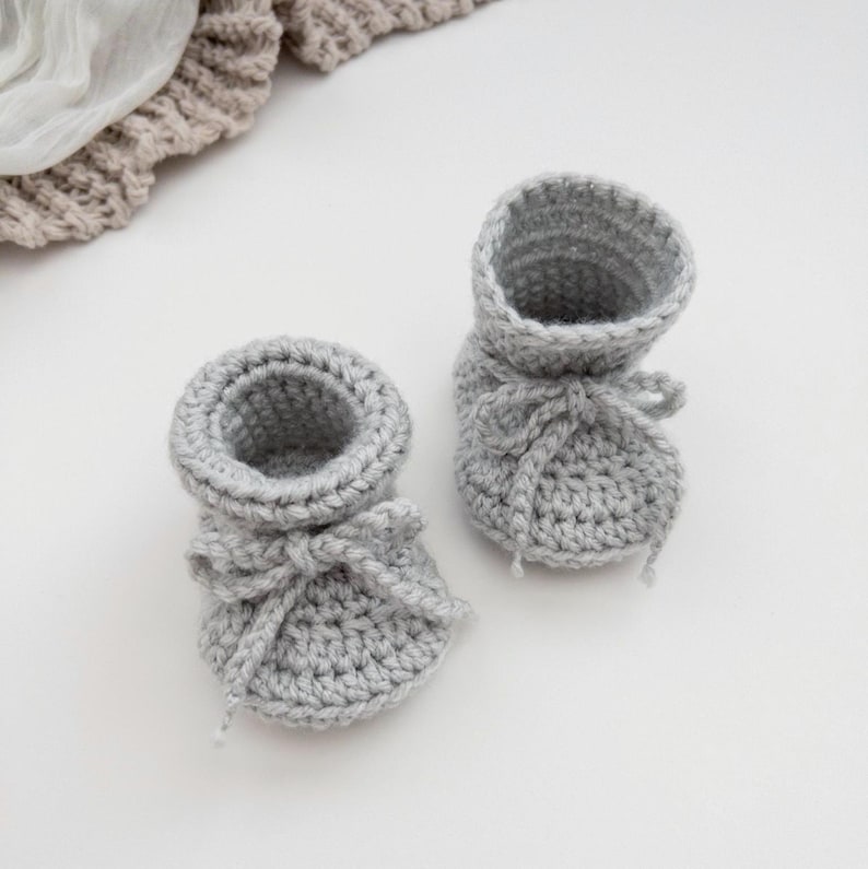 May include: A pair of handmade, light gray crochet baby booties. Each bootie features a rounded toe, a cuff, and a delicate bow at the front. The soft, textured yarn suggests a cozy and comfortable design, perfect for a newborn.