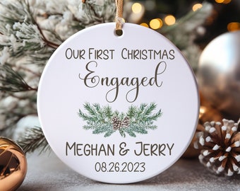 Puede incluir: Adorno navideño de cerámica blanca con cinta dorada. El adorno dice "Our First Christmas Engaged" con los nombres Meghan & Jerry y la fecha 08.26.2023. El adorno tiene un diseño de piñas y agujas de pino verdes y marrones.