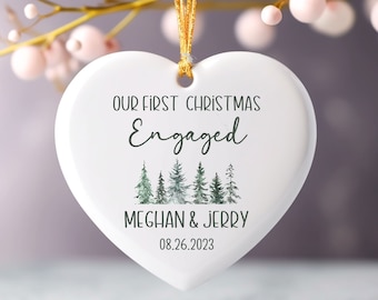 Puede incluir: Adorno navideño de cerámica blanco en forma de corazón con el texto "Our First Christmas Engaged Meghan & Jerry 08.26.2023" y una ilustración de acuarela de un bosque.