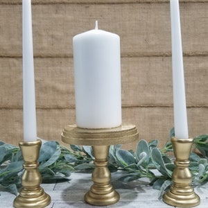 May include: A set of three gold candle holders with white candles. The center candle holder is taller and has a round base. The two side candle holders are shorter and have a round base.