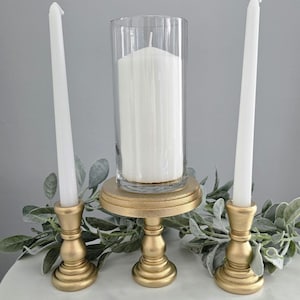 May include: A decorative arrangement featuring three candle holders. Two gold-colored candlestick holders flank a central gold-colored pedestal holding a clear glass cylinder with a white pillar candle. Greenery accents the display.