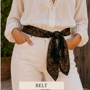 May include: A dark green belt with a gold paisley design is tied around the waist. The belt is worn over white pants and a white blouse. The word "BELT" is displayed in a white rectangle at the bottom of the image.