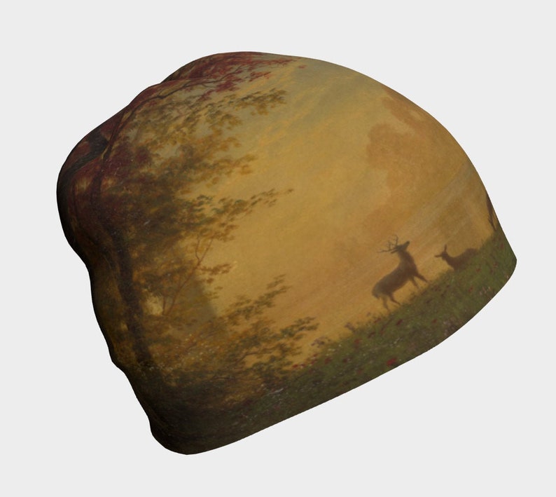 May include: A brown and beige beanie featuring a painting of a woodland scene with two deer in the foreground. The sky is a pale yellow with a touch of orange.