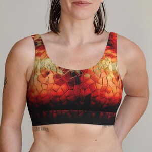 May include: A sports bra with a geometric pattern in shades of red, orange, and gold. The bra has a black band at the bottom and a scoop neckline. The words "Clean Hands" are visible on the skin below the bra.