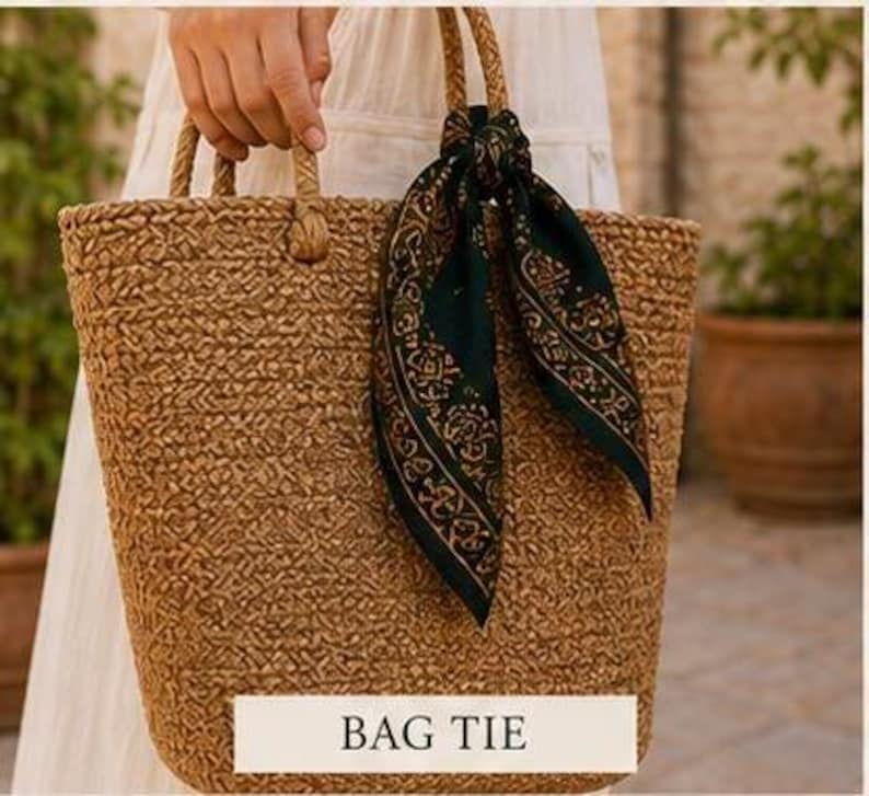 May include: A woven brown tote bag with rope handles. A dark green scarf with a gold pattern is tied to one of the handles. The text "BAG TIE" is displayed on a white rectangle at the bottom of the bag.
