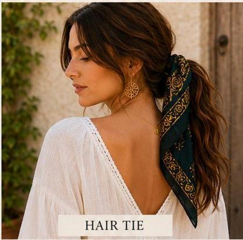 May include: A woman with long brown hair styled in a ponytail, secured with a dark green and gold patterned hair tie. She wears a white top and gold earrings. The text "HAIR TIE" is at the bottom.