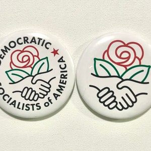May include: Two white round buttons with red, green and black designs. The left button has the text "Democratic Socialists of America" and a red rose with green leaves. The right button has a red rose with green leaves and two hands shaking.