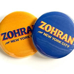 May include: Two round campaign buttons. One is yellow with the text "ZOHRAN FOR NEW YORK CITY" in blue and red. The other button is blue with the same text in yellow and red. The buttons are likely for a political campaign.