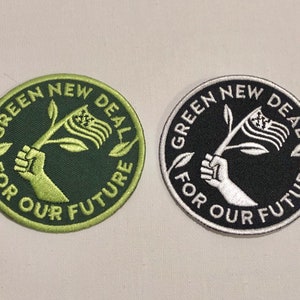 May include: Two embroidered patches with the text "Green New Deal For Our Future". One patch is green on a black background, and the other is white on a black background. Both patches feature a stylized image of a hand holding a branch with a leaf and a flag.